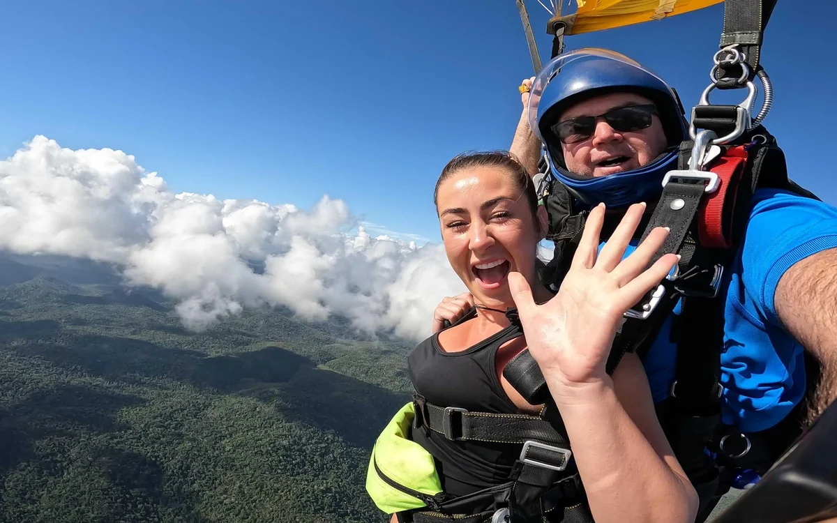 A tandem pair under an open canopy above forested hills and a bank of cloud